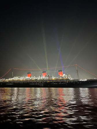 Illuminated vintage ocean liner at night with three red smokestacks, colorful searchlights and string lights reflecting on calm harbor water