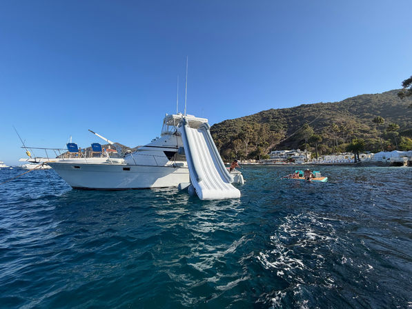 White motor yacht anchored in a sunny coastal cove with a large inflatable water slide into deep blue water, a nearby floating platform with people, and a tree-covered hillside and beach front in the background.