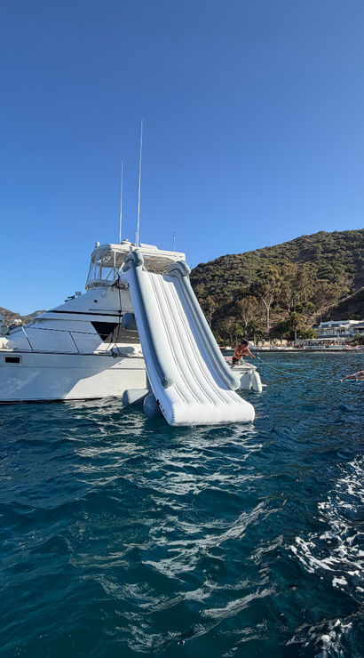 White motor yacht anchored in a sunny coastal cove with a large inflatable water slide into deep blue water, a nearby floating platform with people, and a tree-covered hillside and beach front in the background.