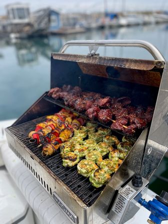 Close-up of a boat-side gas grill at a marina cooking juicy marinated beef skewers, colorful vegetable kebabs, and herb-crusted grilled shrimp with calm harbor water blurred in the background.