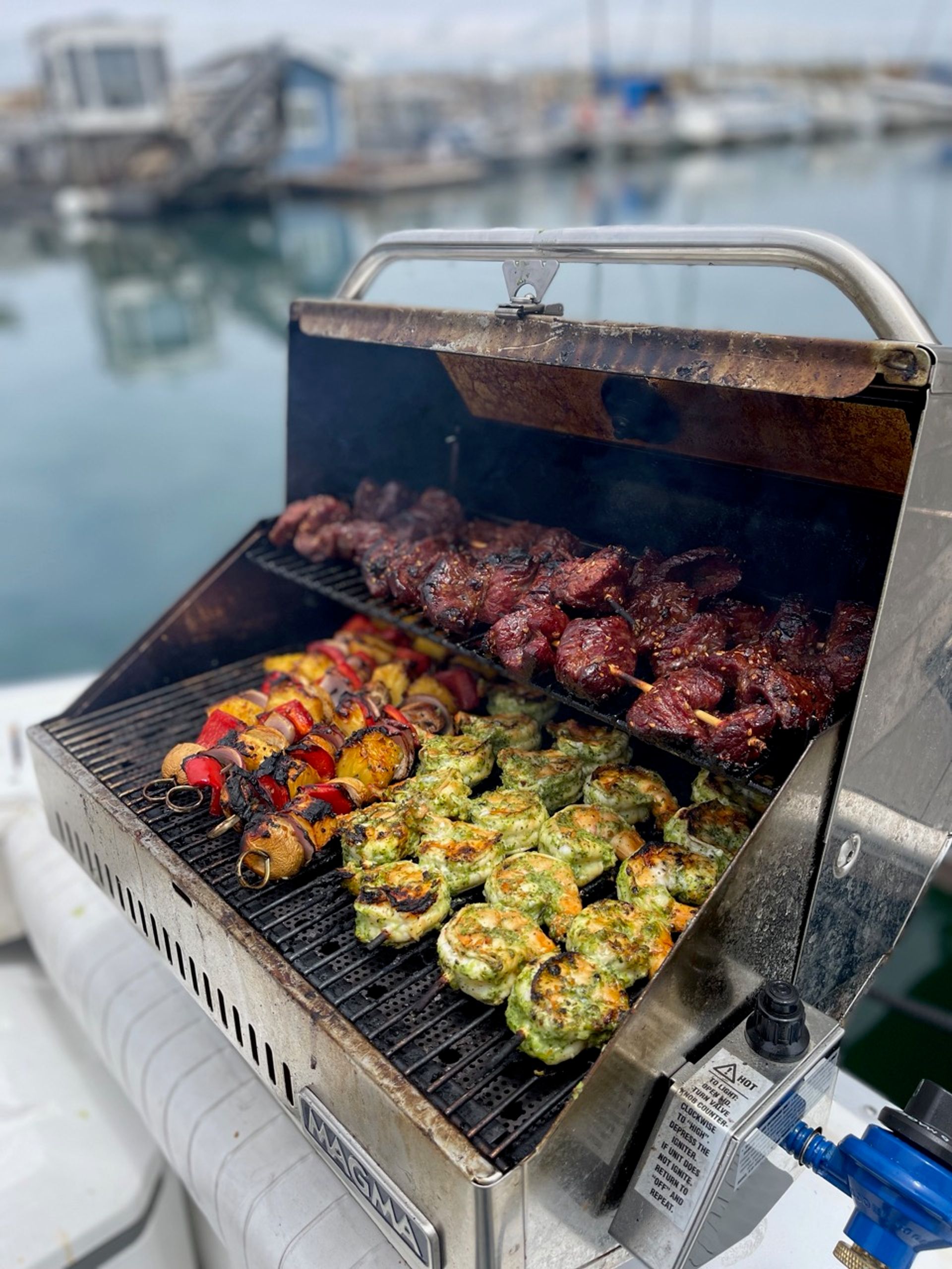 Close-up of a boat-side gas grill at a marina cooking juicy marinated beef skewers, colorful vegetable kebabs, and herb-crusted grilled shrimp with calm harbor water blurred in the background.