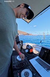 DJ wearing sunglasses and a cap mixing on a controller and laptop aboard a small boat, blue ocean with anchored sailboats and a hilly coastline under a sunny sky