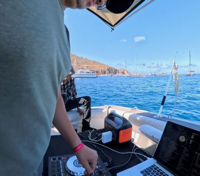 DJ wearing sunglasses and a cap mixing on a controller and laptop aboard a small boat, blue ocean with anchored sailboats and a hilly coastline under a sunny sky