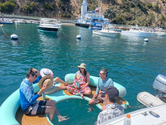 Friends relaxing on a turquoise inflatable boat with a circular swim well in clear blue-green harbor waters, anchored motorboats and hillside buildings on a sunny coastal day