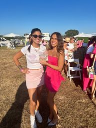 Two smiling women in pink outfits pose in a sunny grassy field at an outdoor daytime event with white tents and folding chairs; one wears sunglasses, a white tee, pink mini skirt and sneakers, the other a bright pink strapless dress and sparkly heels holding a small clear award.