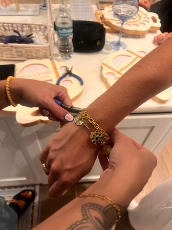 Close-up of hands using pliers to fasten a gold charm bracelet at a hands-on jewelry-making workshop counter, with tools, crafting trays, a water bottle and a wine glass in the background.