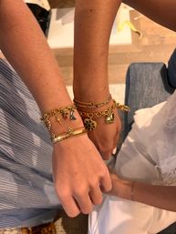Close-up of two wrists wearing stacked gold charm bracelets and slender bangles with floral, star and heart charms, styled over striped and white summer fabrics — fashion wrist jewelry close-up.