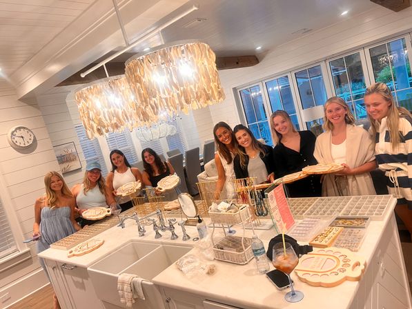 Group of friends at a coastal-style beach house kitchen island enjoying a DIY jewelry-making party with bead displays, wooden trays, shell chandeliers, and bright white shiplap interior