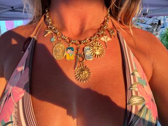 Close-up of a gold charm necklace with sun medallions and colorful charms on a tanned chest, worn with a floral halter bikini in warm sunset beach light.