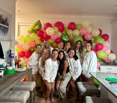 Smiling group of friends posing in a home kitchen in front of a pink, lime-green and cream balloon arch with citrus foil balloons and a casual party table setup.