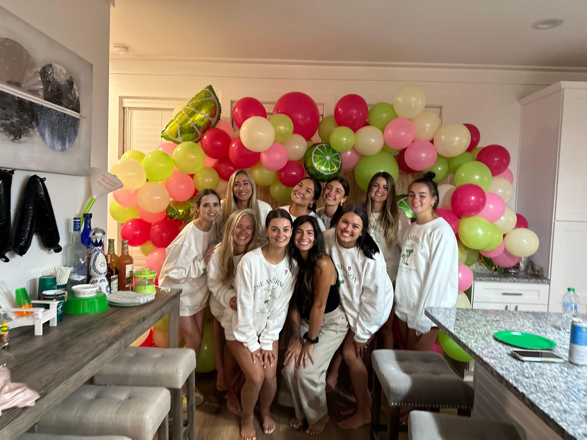 Smiling group of friends posing in a home kitchen in front of a pink, lime-green and cream balloon arch with citrus foil balloons and a casual party table setup.