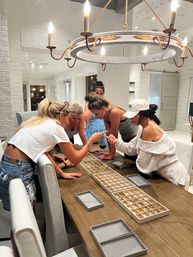 Group of five women leaning over a long wooden dining table in a bright white farmhouse-style kitchen, examining jewelry trays under a rustic chandelier.