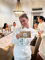 Smiling woman holds up a gold charm necklace card during a girls’ night/bachelorette-style gift reveal as friends in matching white sweatshirts open presents around a dining table under a beaded chandelier.