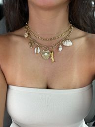 Close-up of a woman’s décolletage wearing layered gold charm necklaces — chunky chains with shell, cross, heart and mermaid-tail pendants over a beige strapless top