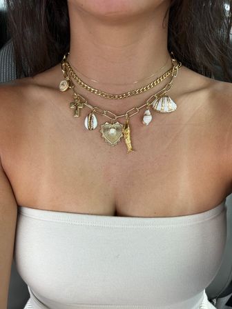 Close-up of a woman’s décolletage wearing layered gold charm necklaces — chunky chains with shell, cross, heart and mermaid-tail pendants over a beige strapless top