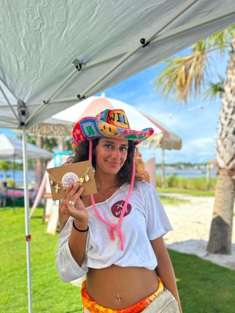Smiling vendor at a beachside outdoor market holding a handmade shell-charm necklace on a kraft card, wearing a colorful crocheted cowboy hat and white crop tee with palm trees and waterfront in the sunny background.