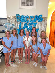 Eight women in matching light-blue pajama sets smiling and posing barefoot indoors in front of a silver fringe backdrop, blue letter balloons and a large diamond-shaped balloon for a bachelorette celebration