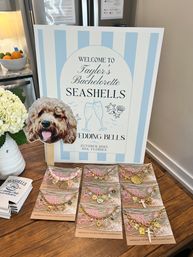 Playful coastal bachelorette display: pale blue-striped welcome sign with seaside wedding theme and location 30A, Florida, dog-face photo prop on a stick, white hydrangea in a vase, and a wooden table spread of gold seashell charm bracelets and koozies.