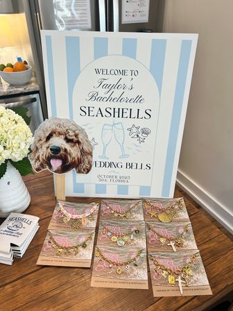 Playful coastal bachelorette display: pale blue-striped welcome sign with seaside wedding theme and location 30A, Florida, dog-face photo prop on a stick, white hydrangea in a vase, and a wooden table spread of gold seashell charm bracelets and koozies.