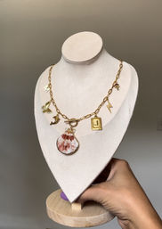 Gold link charm necklace featuring a pink-and-white scallop shell pendant, butterfly, crescent moon, lightning bolt and initial charm, shown on a beige fabric display bust held by a hand against a neutral background.