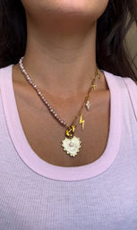 Close-up of neck and collarbone wearing playful layered jewelry: a pink pearl strand paired with a gold link chain holding a textured gold heart pendant, lightning bolt, tiny cross and baroque pearl over a light pink ribbed tank top.