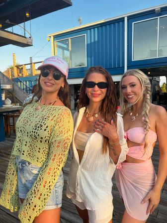 Three sun-kissed women in summer outfits posing on a coastal boardwalk outside a blue waterfront bar at golden hour — crochet top with denim shorts, white coverup, pink bikini wrap, sunglasses and shell necklaces.