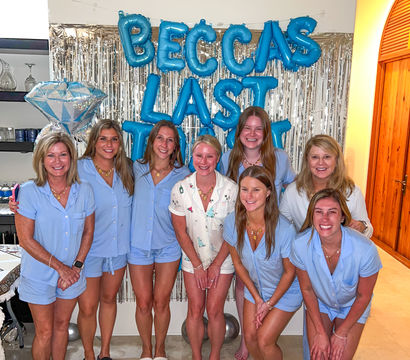 Eight women in matching light-blue pajama sets smiling and posing barefoot indoors in front of a silver fringe backdrop, blue letter balloons and a large diamond-shaped balloon for a bachelorette celebration