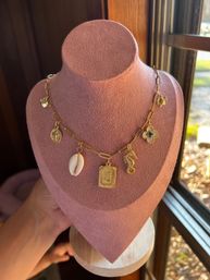 Close-up of a gold paperclip-chain charm necklace — cowrie shell, engraved 'J' rectangle, seahorse, coin and quatrefoil charms — displayed on a pink velvet bust by a sunlit window.
