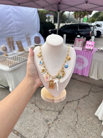 Hand holding a beige necklace bust displaying a gold charm chain necklace with beach-themed charms (shells, palm tree, seahorse, blue enamel) at an outdoor artisan market booth under a pink canopy.