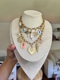 Layered gold charm necklace on cream display bust featuring a jeweled heart locket, pink seahorse plaque, enamel blue fish oval, baroque pearl, shell, star and fish charms — playful statement jewelry