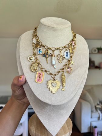 Layered gold charm necklace on cream display bust featuring a jeweled heart locket, pink seahorse plaque, enamel blue fish oval, baroque pearl, shell, star and fish charms — playful statement jewelry