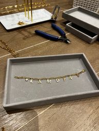 Gold paperclip-chain bracelet with dangling letter charms laid on a gray tray at a jewelry workbench, surrounded by blue-handled wire cutters, stacked display trays and extra gold chains.
