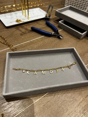 Gold paperclip-chain bracelet with dangling letter charms laid on a gray tray at a jewelry workbench, surrounded by blue-handled wire cutters, stacked display trays and extra gold chains.