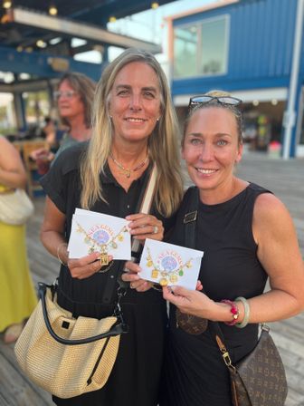 Two smiling women on a coastal wooden boardwalk outside a blue shop, each holding a “Sea Gems” card displaying gold charm bracelets — summer jewelry shopping vibe.