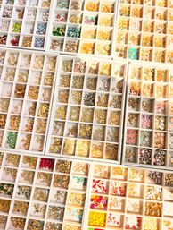 Close-up top-down view of white organizer trays filled with gold-tone charms, colorful beads, enamel accents and jewelry-making supplies for beading and crafts.