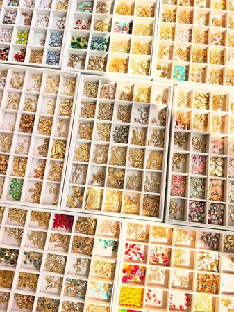 Close-up top-down view of white organizer trays filled with gold-tone charms, colorful beads, enamel accents and jewelry-making supplies for beading and crafts.