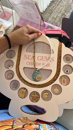 Hand holding a sea-themed charm display board with a pearl-and-chain necklace featuring a blue shell pendant with a gold spiral, surrounded by assorted gold coastal charms (starfish, palm tree, seashell) in a pink organizer on a woven rug.