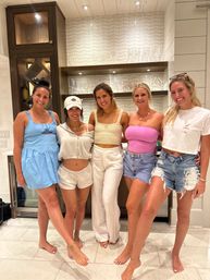 Five smiling women in casual summer outfits pose barefoot in a bright coastal-style kitchen and wet bar with open glassware shelves, shiplap walls, and a wine fridge — fun vacation-home group photo.