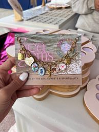Close-up of a hand holding a card showing a sparkly gold charm bracelet with colorful enamel charms—floral ovals, heart, cross and lock—on a sand-and-sea themed backing at an outdoor craft market