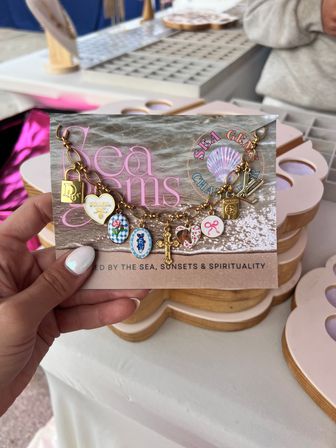 Close-up of a hand holding a card showing a sparkly gold charm bracelet with colorful enamel charms—floral ovals, heart, cross and lock—on a sand-and-sea themed backing at an outdoor craft market