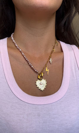 Close-up of a neckline in a light pink ribbed tank top showcasing stylish layered necklaces: a pink freshwater pearl strand with a gold heart pendant featuring an opal center, and a gold link chain with lightning, cross and baroque pearl charms.