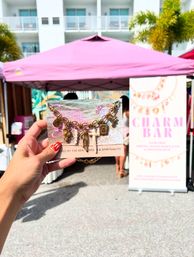 Hand holding a beach-inspired gold charm bracelet on a display card in front of a pink vendor tent and palm trees at a sunny outdoor craft market.