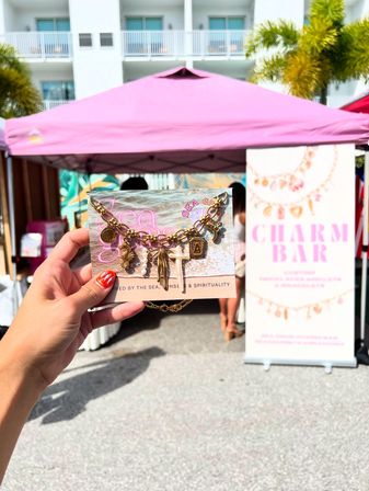 Hand holding a beach-inspired gold charm bracelet on a display card in front of a pink vendor tent and palm trees at a sunny outdoor craft market.