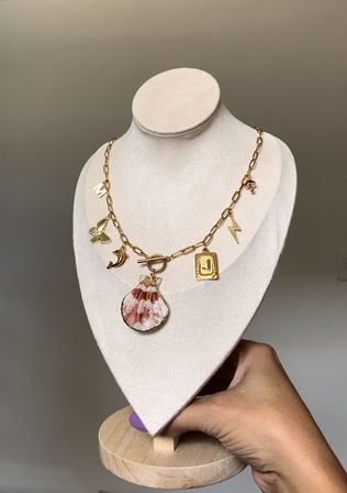 Gold charm necklace with pink-and-white scallop shell pendant and assorted charms (moon, clover, lightning bolt, letter tag) displayed on a beige velvet jewelry bust with wooden base held by hand