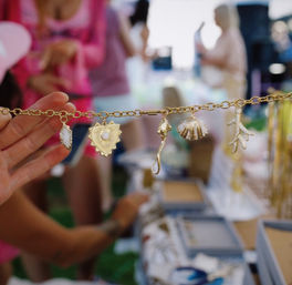 Close-up of a gold charm bracelet with heart, shell, seahorse and coral charms held up in front of an outdoor market stall