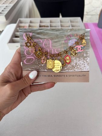 Hand holding a seaside display card at a beach market showing a gold charm bracelet with mixed charms — coin tag, pink enamel heart, Hamsa hand, shell and an 'N' initial.