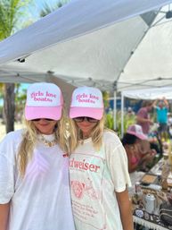 Two women in matching pink-and-white 'girls love boats' trucker hats and sunglasses under a white canopy at a sunny beachside outdoor market, one wearing a vintage-style beer tee.