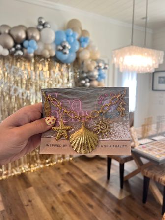 Hand holding a gold sea-themed charm bracelet on packaging—starfish, scallop shell, sun and seahorse charms—against a blurred gold fringe and blue-and-cream balloon party backdrop.