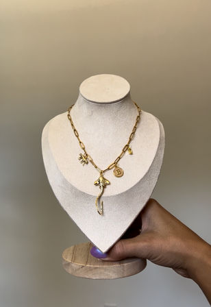 Gold paperclip-chain charm necklace with ocean-inspired stingray pendant and round 'B' initial charm, displayed on a beige velvet necklace bust held by a hand — product jewelry photo.