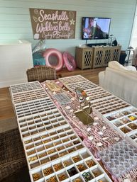 Coastal-themed bachelorette craft setup in a beach house living room with a “Seashells and Wedding Bells” sign, pink pool floats, TV and a large DIY charm bar of organized trays holding gold charms, shells, beads and a tabletop mirror.